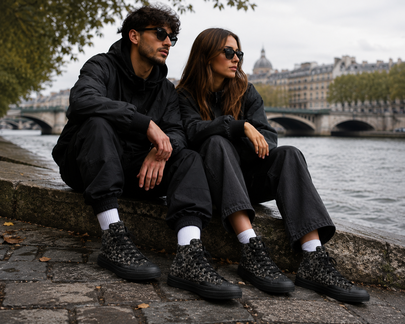 Couple portant des Converse Chuck 70 Marquis Hi Patta Black Mineral Gray Rosin assis au bord d'une rivière urbaine, tenue streetwear noire, sneakers montantes camouflage noir et gris, ambiance lifestyle premium, pont et architecture citadine en arrière-plan.